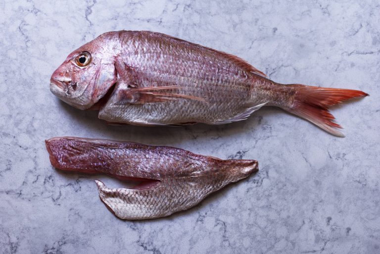 A whole raw Australian snapper with two skin-on fillets laid beside it on a white marble surface, showing the yield from a single fish ready for pan searing.