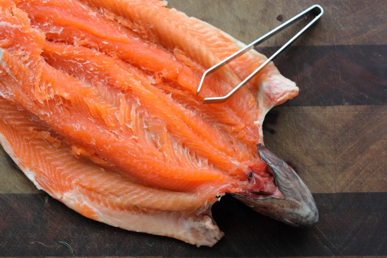 A whole raw rainbow trout laid on a dark wooden cutting board, surrounded by fresh dill, garlic cloves, lemon, and tomato