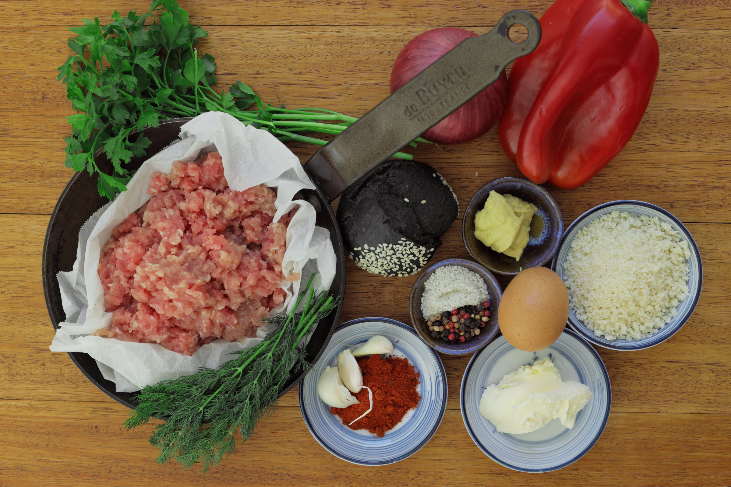 Mise en place for GEOFF's Sustainable Carp Burger: minced carp fillet in a baking-paper-lined bowl, flat-leaf parsley and dill, red onion, red capsicum, smoked paprika, garlic cloves, egg, Dijon mustard, sea salt and mixed peppercorns, panko breadcrumbs and sour cream, arranged on a timber board with a de Buyer mezzaluna knife.