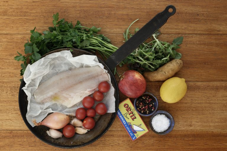 Cast iron pan holding a skinless barramundi fillet on baking paper, surrounded by cherry tomatoes, shallots, and garlic cloves. Alongside: a tin of Ortiz anchovies, fresh flat-leaf parsley, mixed salad greens, a pomegranate, a potato, a lemon, and small bowls of sea salt flakes and mixed peppercorns, arranged on a timber surface.