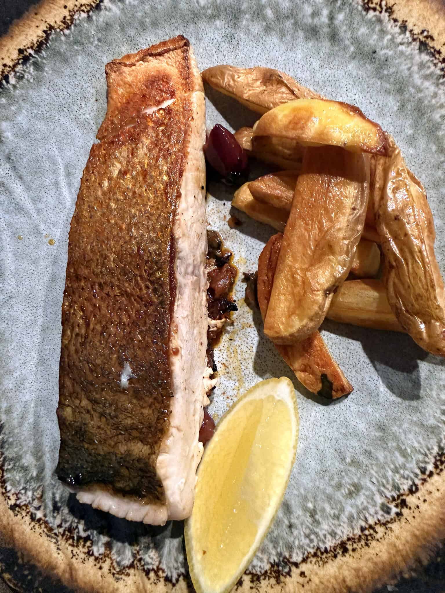 A skin-on kingfish fillet pan-seared to a deep golden brown, resting on a Provençal sauce with Kalamata olives and capers, served alongside roasted skin-on potato wedges and a lemon wedge on a speckled ceramic plate.