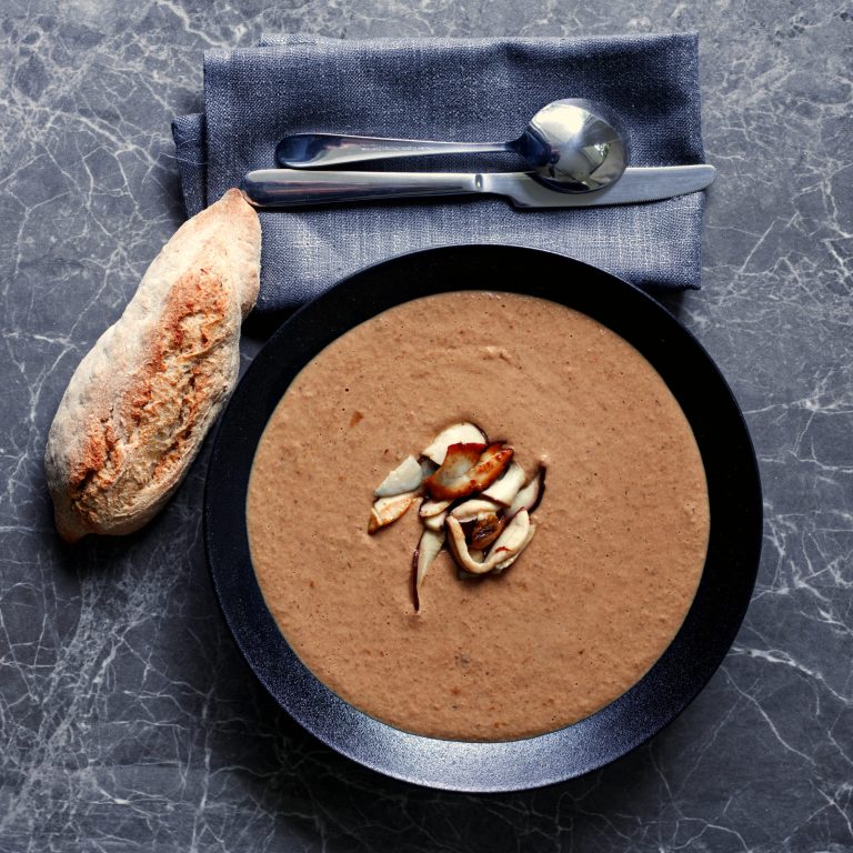 A wide dark bowl filled with smooth brown porcini soup, garnished at the centre with a small pile of sautéed porcini slices. A sourdough roll sits to the left of the bowl. A folded blue linen napkin with two silver spoons rests at the top of the frame, on a grey marble surface.