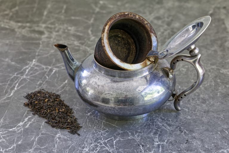 A well-worn silver teapot with its hinged lid open and mesh strainer basket lifted, resting on a grey marble surface. A small pile of loose black tea leaves sits to the left. The pot shows decades of use — patina on the exterior, deep staining inside the strainer. Shot from slightly above at an angle.