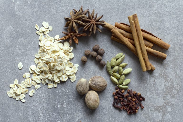 verhead flat-lay of chai spice porridge ingredients on grey concrete. Rolled oats, star anise, cinnamon sticks, cardamom pods, nutmeg, allspice berries and cloves arranged in groups.
