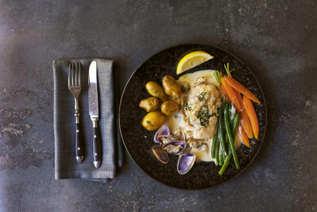 Overhead wide view of poached bearded rock cod in cream sauce with Goolwa pipis, roasted baby potatoes, green beans and baby carrots on a dark ceramic plate, with grey linen napkin and silver cutlery alongside on a grey concrete surface