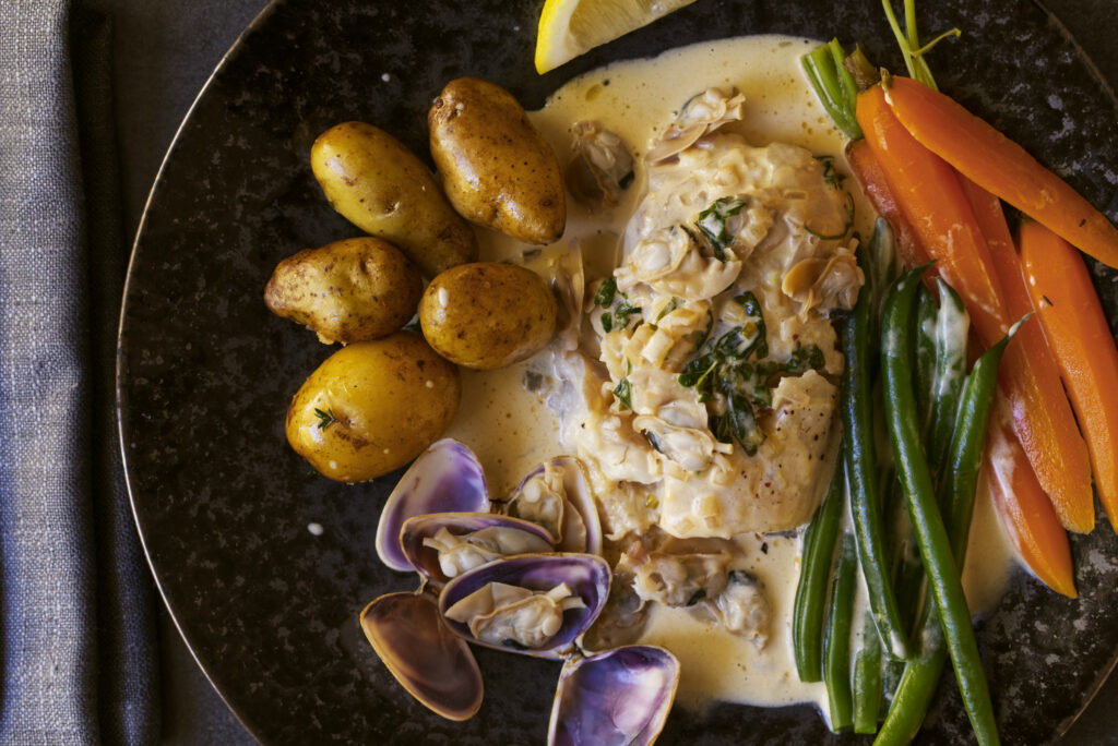 Overhead view of poached bearded rock cod in cream sauce with Goolwa pipis, roasted baby potatoes, green beans and baby carrots on a dark ceramic plate with grey linen