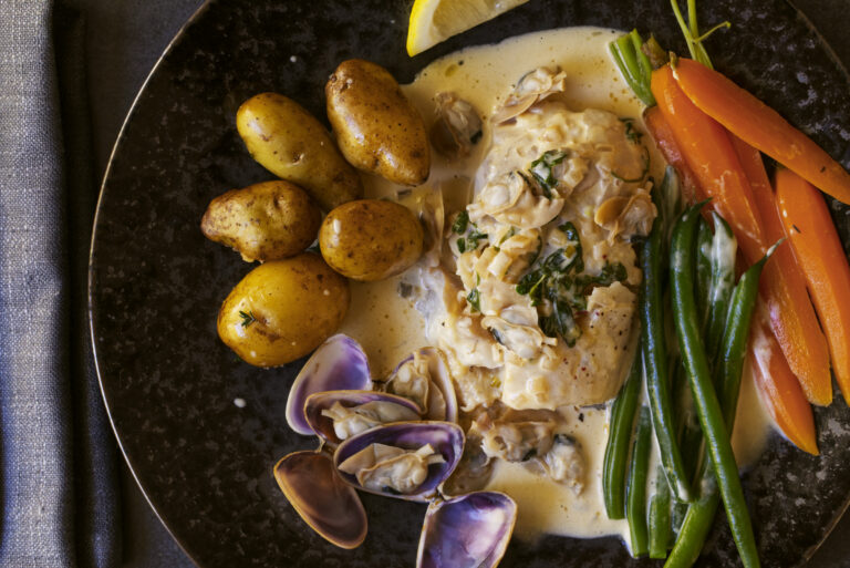 Overhead view of poached bearded rock cod in cream sauce with Goolwa pipis, roasted baby potatoes, green beans and baby carrots on a dark ceramic plate with grey linen
