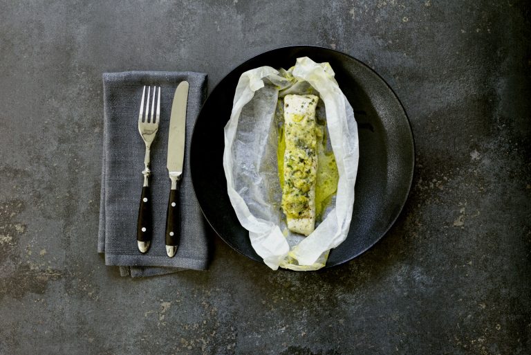 Overhead view of an opened white baking paper parcel on a black plate, with a centre-cut white fish fillet sitting in pooled golden urchin butter. Chopped herbs and desert lime pieces are visible on top of the fillet. Grey linen napkin and cutlery to the left on a concrete-grey surface.