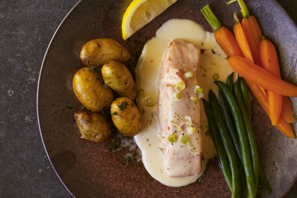 Overhead view of poached Ora King salmon fillet with creamy velout&eacute; sauce, roasted new potatoes, green beans and baby carrots on a dark ceramic plate against grey concrete.