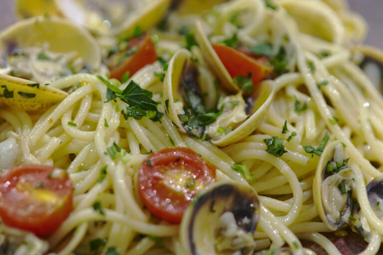 Macro close-up of cooked spaghetti alle vongole, showing open clam shells, halved cherry tomatoes and chopped parsley glistening in olive oil.