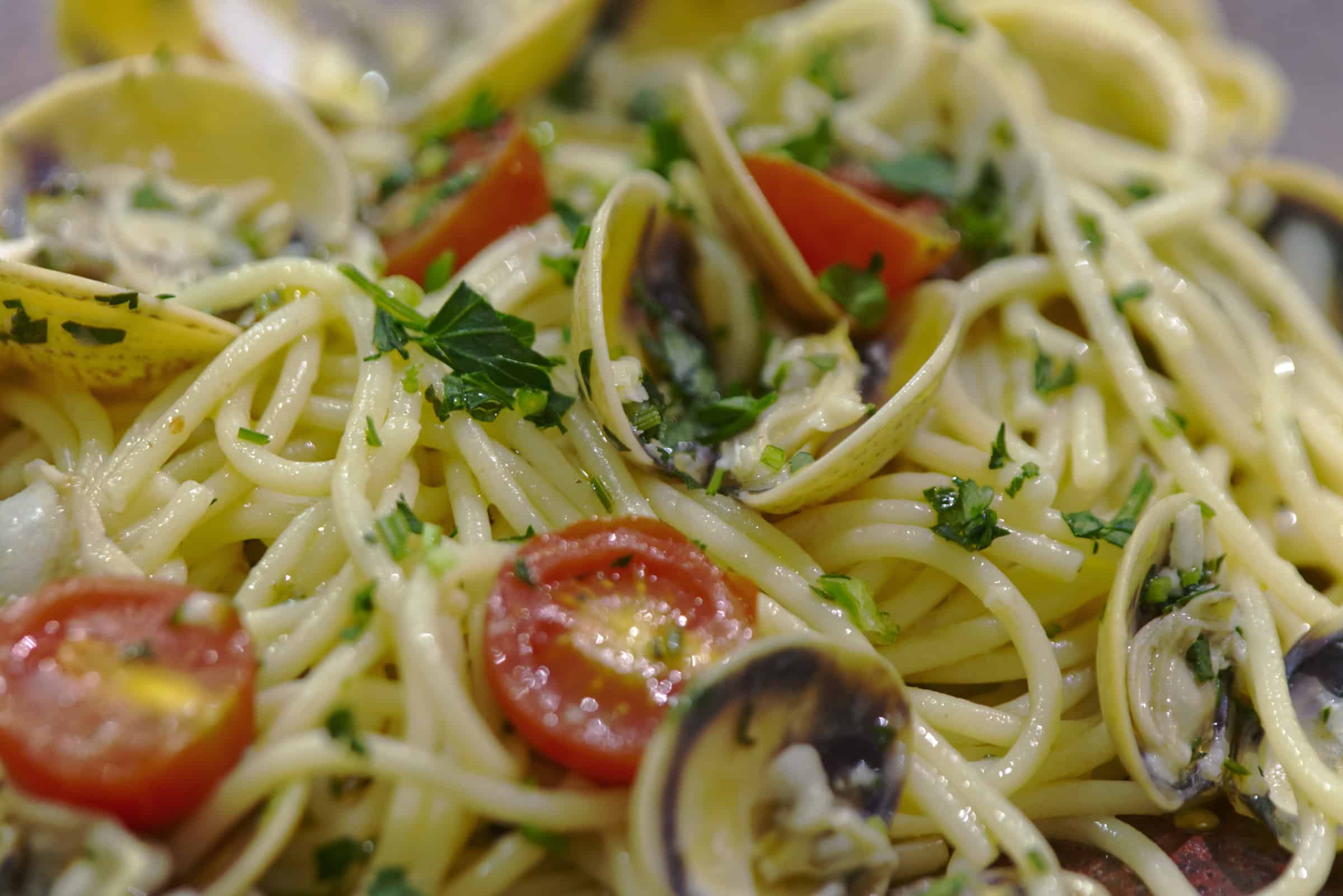 Macro close-up of cooked spaghetti alle vongole, showing open clam shells, halved cherry tomatoes and chopped parsley glistening in olive oil.