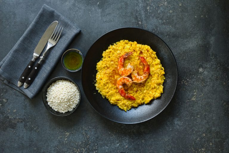 Overhead view of saffron risotto with tiger prawns in a black bowl on dark metallic surface, with a small bowl of arborio rice and sauce dish alongside grey linen and cutlery