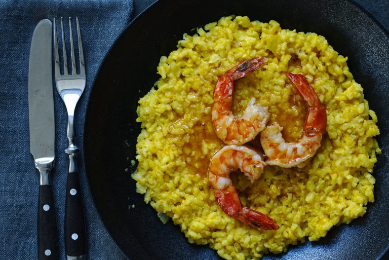 Overhead close crop of saffron risotto topped with three tiger prawns in bisque sauce, served in a black bowl on a dark metallic surface with grey linen and cutlery