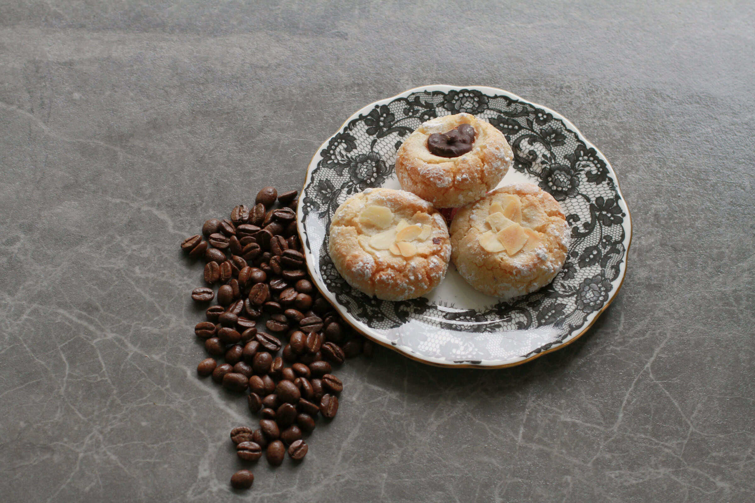 Three round amaretti biscuits with a golden, crackled surface dusted in icing sugar, sitting on a white plate with a black lace border. One biscuit is topped with a dark chocolate button, the other two with flaked almonds. Coffee beans are scattered on the stone surface beside the plate.