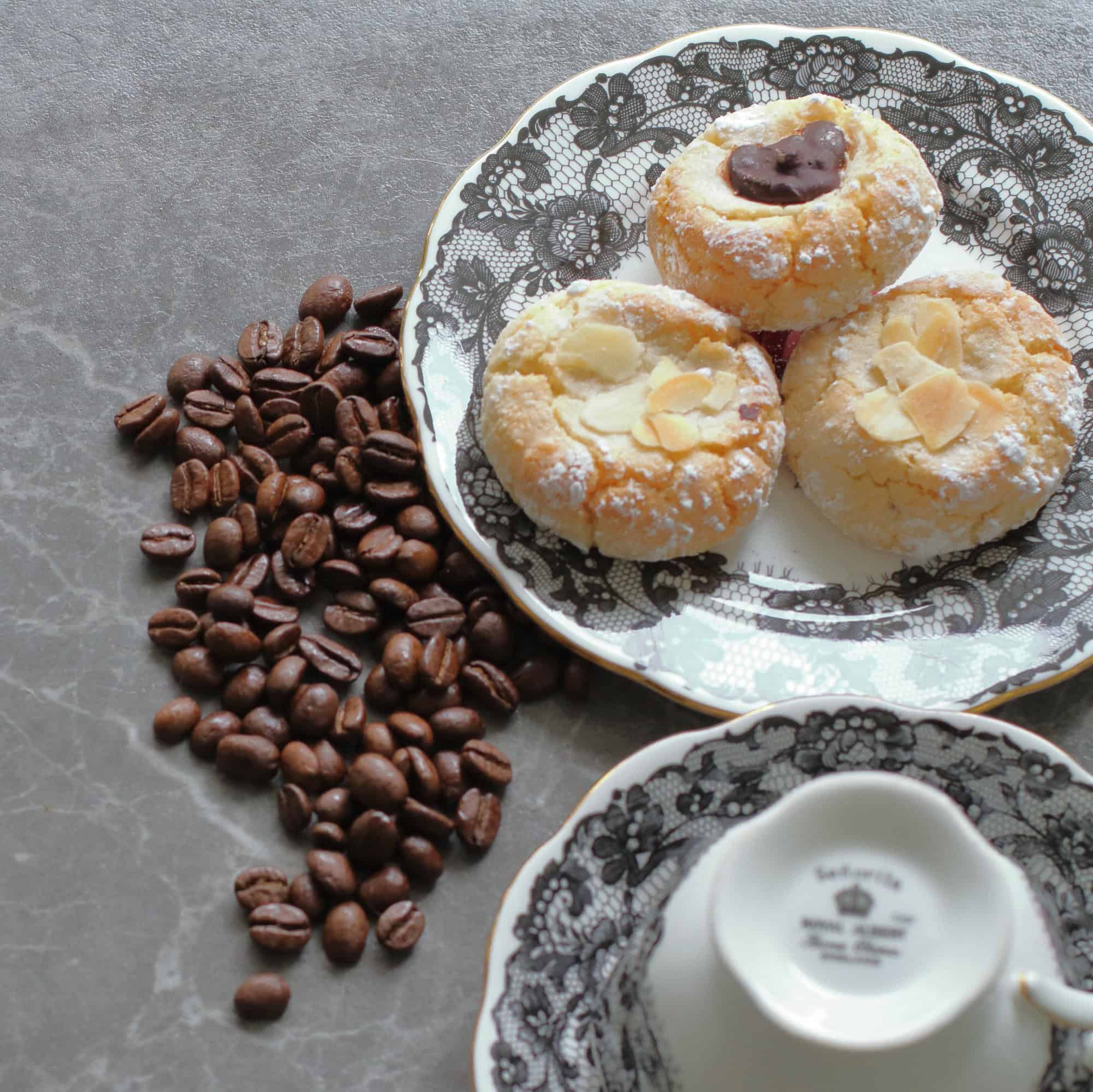 A close crop of three golden amaretti biscuits dusted with icing sugar on a black lace-patterned plate, one topped with a dark chocolate button and two with flaked almonds, coffee beans scattered to the left, with an upturned bone china cup and saucer showing a maker's mark on the base in the foreground.