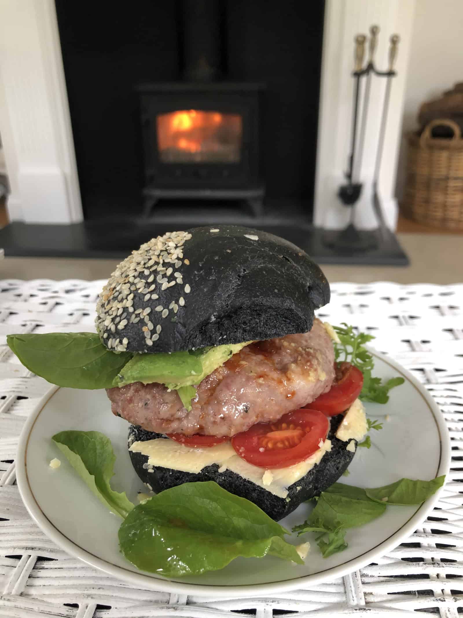 A split charcoal brioche roll filled with a lamb patty, sliced tomato, cheese, avocado and lettuce, served on a white plate.