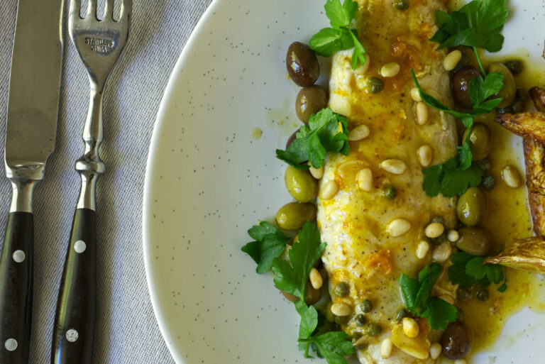 Close-up of pan-cooked fish fillet in citrus and olive sauce, garnished with pine nuts, capers, and parsley, with the edge of a knife and fork resting on a linen napkin.