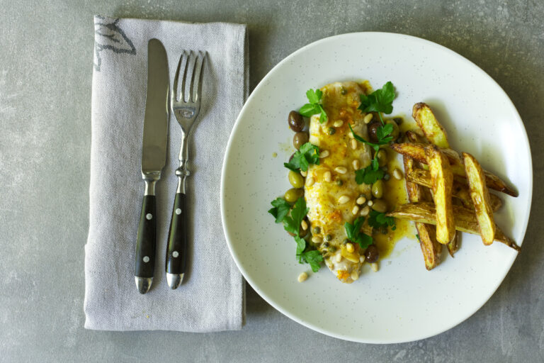 Overhead shot of a pan-cooked fish fillet topped with olives, capers, pine nuts and parsley in a citrus sauce, served with golden roasted potato wedges on a white plate, cutlery and a linen napkin alongside, on grey concrete.