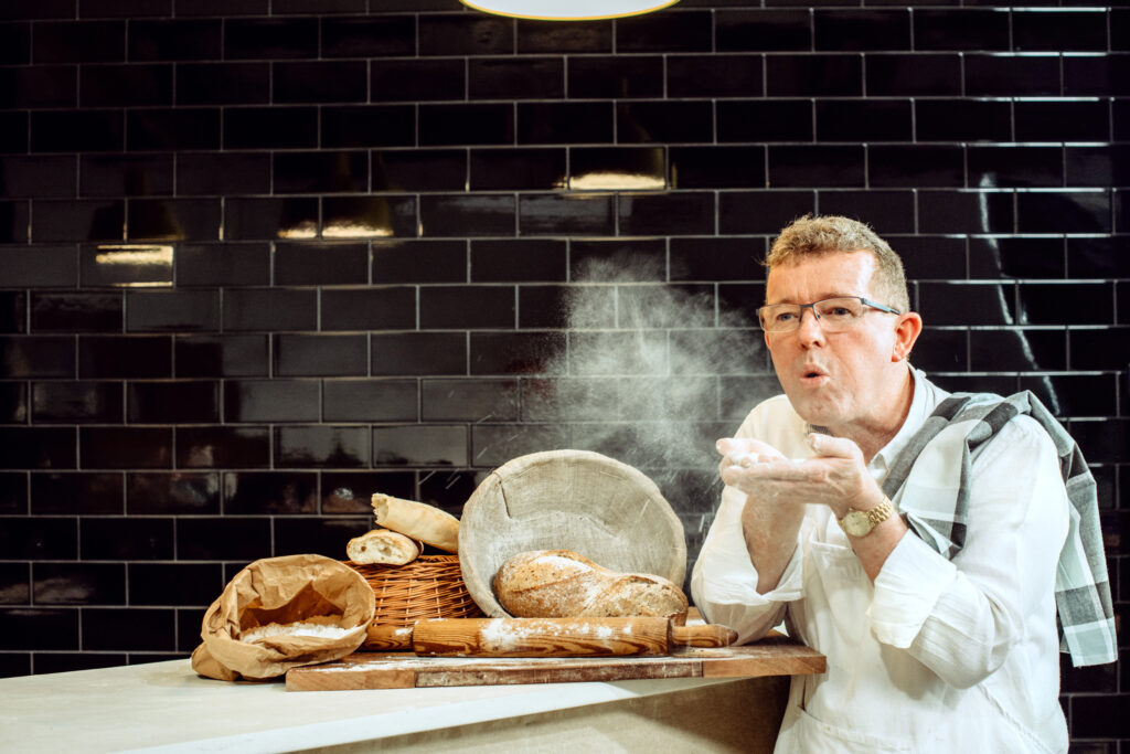 Geoff mid-motion, tossing flour over a board of fresh bread, baskets and rolling pins