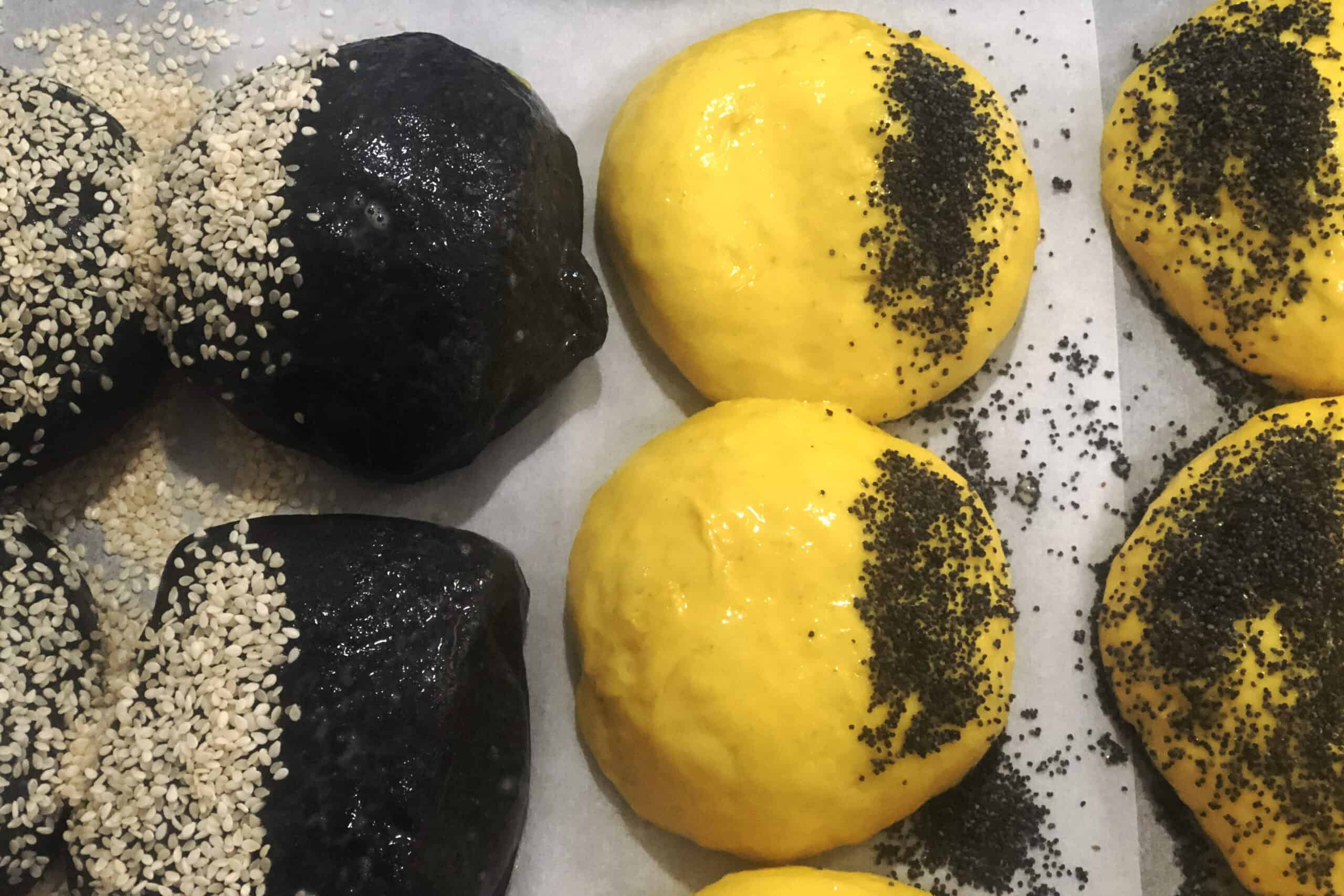 Close-up of charcoal and turmeric brioche rolls with egg glaze brushed over half of each surface, seeds scattered nearby ready to be pressed on.