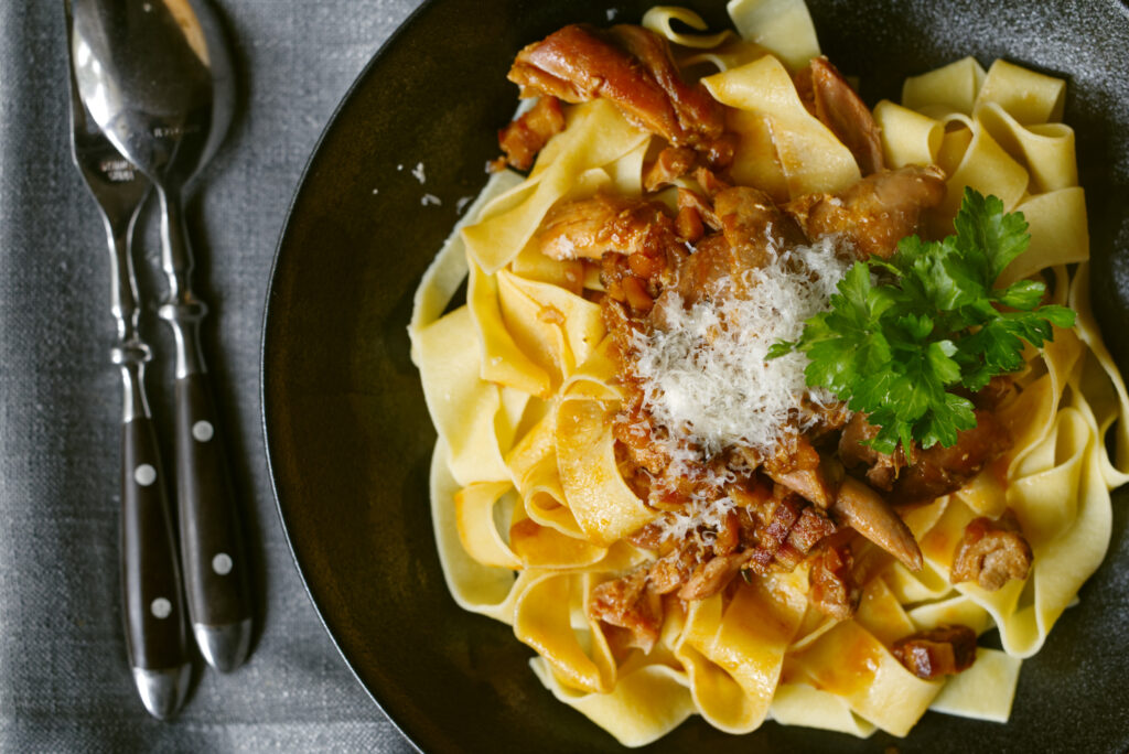 Close overhead shot of a dark plate showing wide pappardelle ribbons topped with shredded wild rabbit rag&ugrave;, a mound of finely grated Parmigiano Reggiano, and flat leaf parsley leaves, with a grey linen napkin and cutlery to the left.