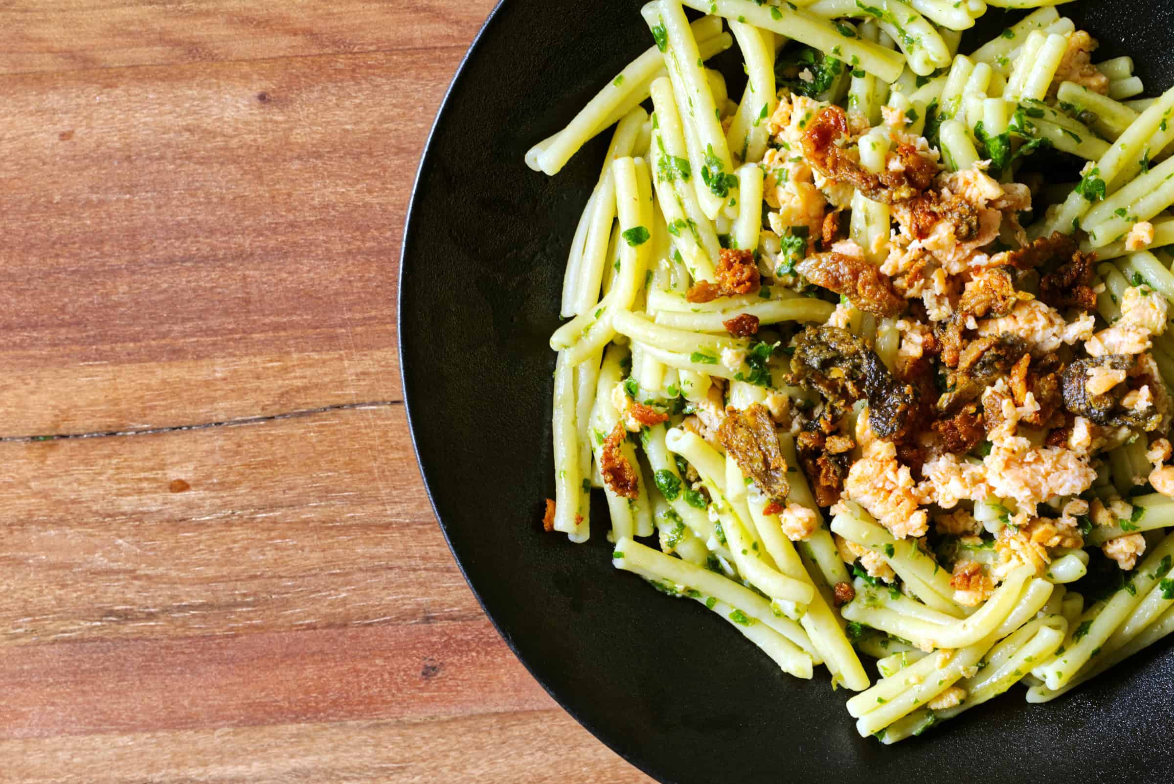 Close-up overhead view of Casareccia pasta coated in a bright green herb sauce on a dark round plate, topped with finely minced salmon and pieces of deep golden crispy salmon skin scattered across the surface.
