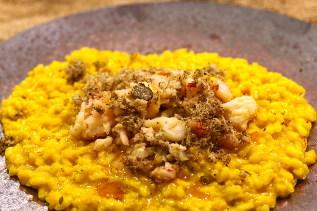 A close-up of saffron-yellow risotto on a dark plate, topped with pieces of Balmain bug meat, grated truffle, and a single visible truffle shaving, with a little butter pooling at the edge.