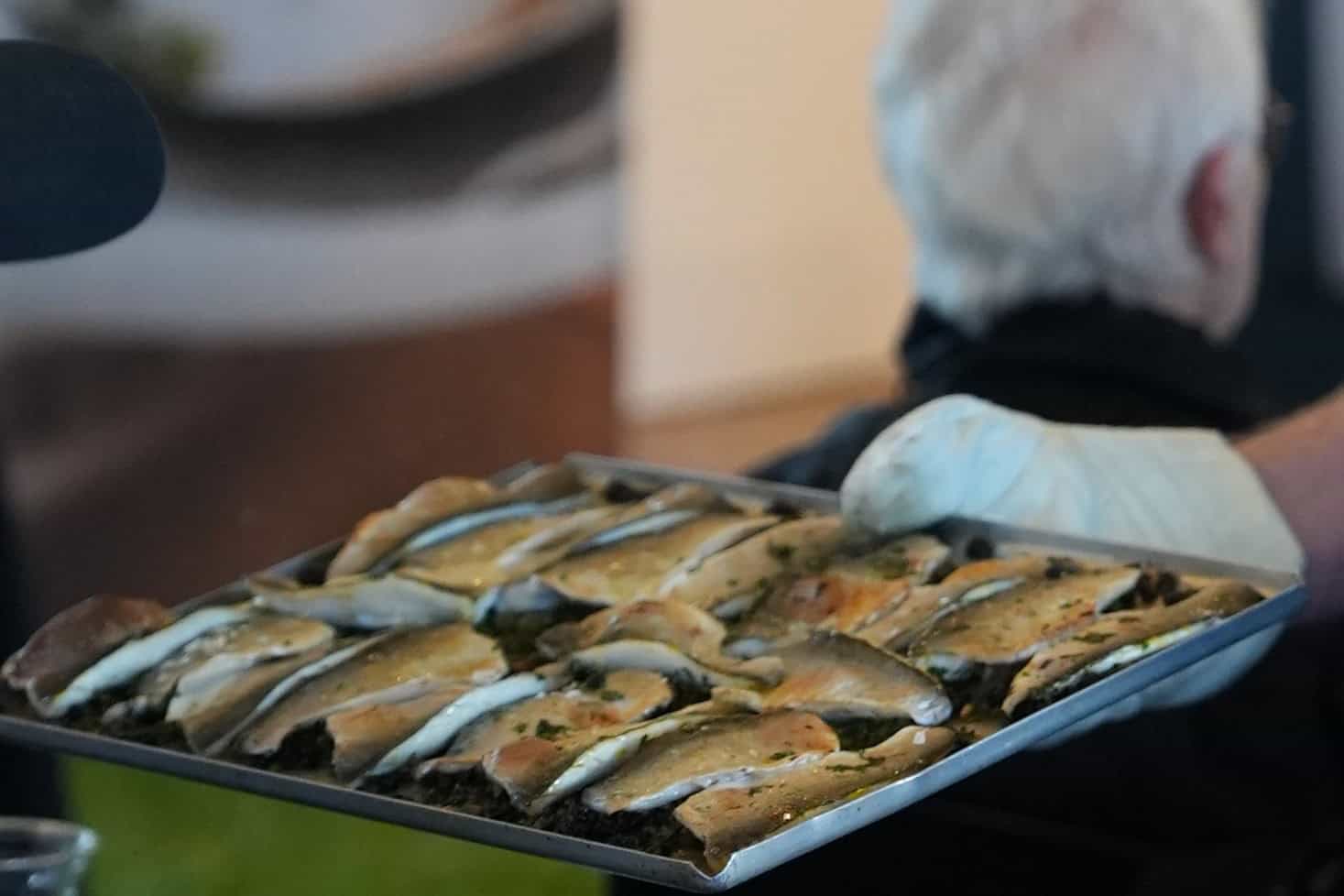 A metal tray of butterflied Coorong mullet fillets arranged skin-side up in overlapping rows over a dark herb stuffing, glistening with oil, held at an angle by a gloved hand.
