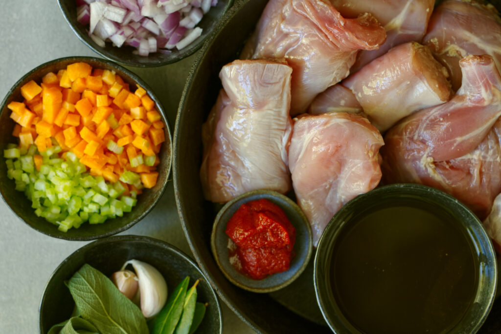 Overhead shot of raw wild rabbit pieces and mirepoix ingredients arranged in dark bowls on a grey surface, including diced red onion, diced carrot and celery, tomato paste, garlic cloves, sage leaves, bay leaves, and a bowl of dark veal stock.