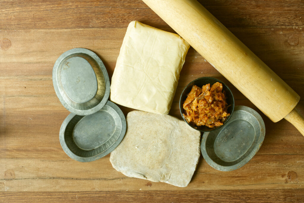 Overhead shot on a dark timber board showing a block of puff pastry, a slab of wholegrain spelt shortcrust pastry, a small dark bowl of shredded wild rabbit rag&ugrave;, three oval tin pie dishes, and a wooden rolling pin.