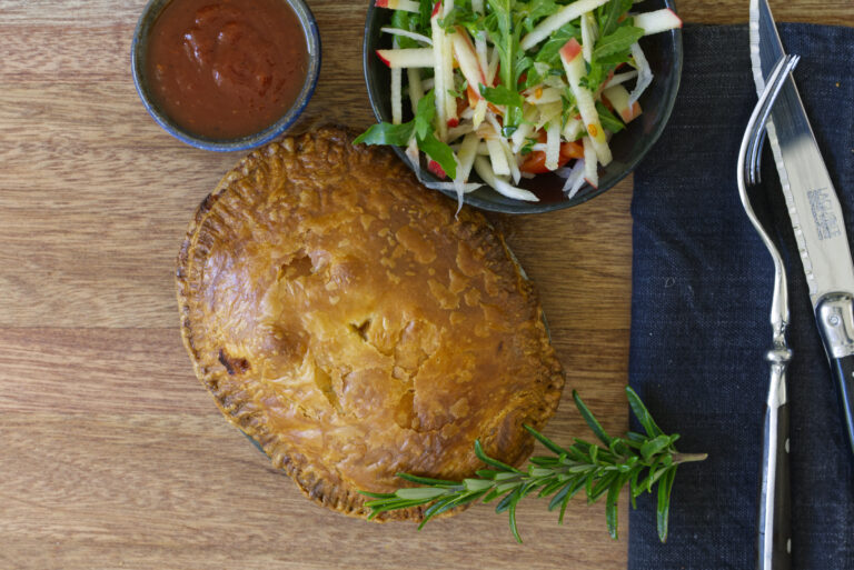 Overhead shot of a baked wild rabbit ragù pie in an oval tin on a dark timber board with a navy linen napkin, a sprig of rosemary across the pie, a bowl of fennel, rocket, crispy apple and tomato salad, a small bowl of tomato sauce, and cutlery to the right.