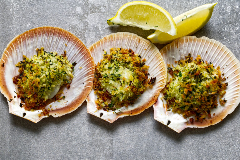 Three scallops in their shells topped with a golden herb and garlic breadcrumb crust, garnished with lemon wedges on a grey concrete surface.