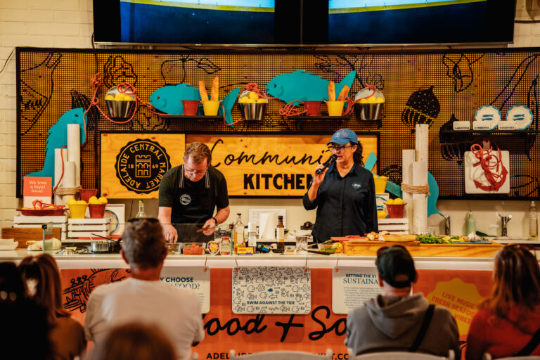 A wide shot of the Community Kitchen stage at Adelaide Central Market, with a man in an apron preparing food at the bench and a woman holding a microphone speaking to a seated audience, framed by branded signage, buckets of lemons and cutout fish decorations.